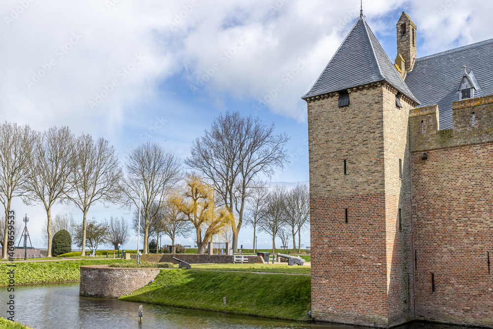 Obraz premium Bare trees, moat, green grass, Radboud castle tower with brick walls and canons in the background, sunny day with a blue sky and white clouds in Medemblik, Noord-Holland, Netherlands