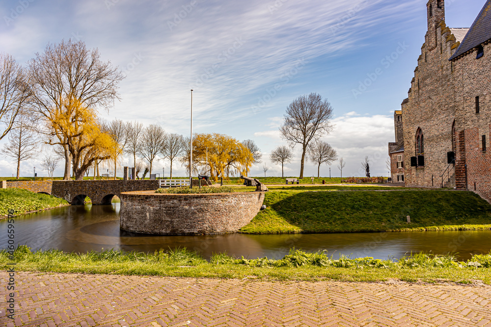 Moat with its bridge, cannons and bare trees in the background, partial view of Radboud castle, sunny day with a blue sky and white clouds in Medemblik, Noord-Holland, Netherlands. Long exposure photo