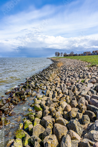 Water breaking on the stones on the shoreline at the shore of Lake Ĳsselmeer, horizon, house and bare trees in the background, cloudy day with stormy clouds, in Medemblik, Noord-Holland, Netherlands