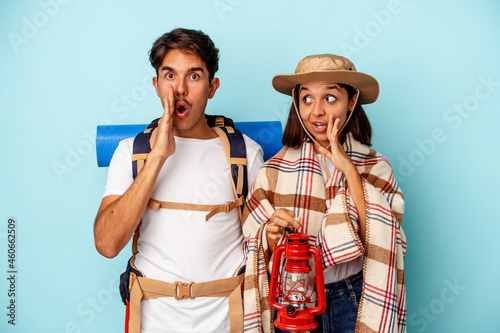 Young mixed race hiker couple isolated on blue background is saying a secret hot braking news and looking aside