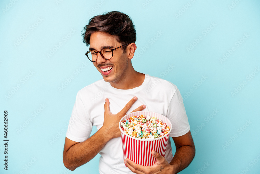 Young mixed race man eating popcorns isolated on blue background laughing and having fun. Young mixed race man eating popcorns isolated on blue background laughing and having fun.