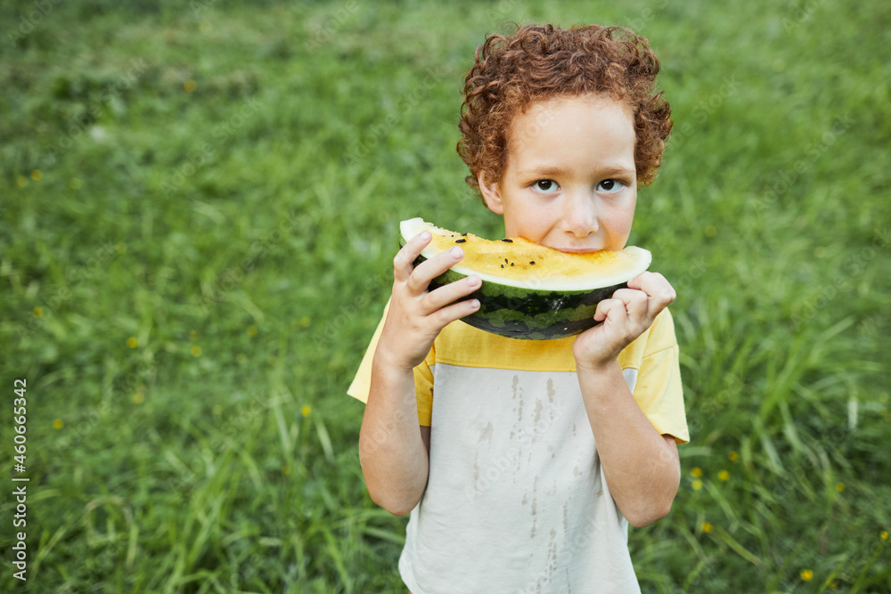 Waist up portrait of curly boy eating watermelon outdoors and looking at camera Waist up portrait of curly boy eating watermelon outdoors and looking at camera