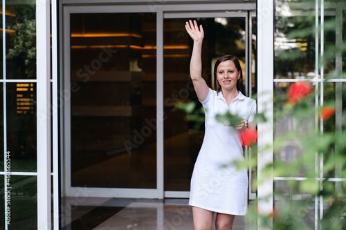 an adult beautiful woman in a white dress waves to someone, a neighbor or a guest client of a cafe, standing in the doorway. a friendly smile on her face, summer and flowers. meeting or parting