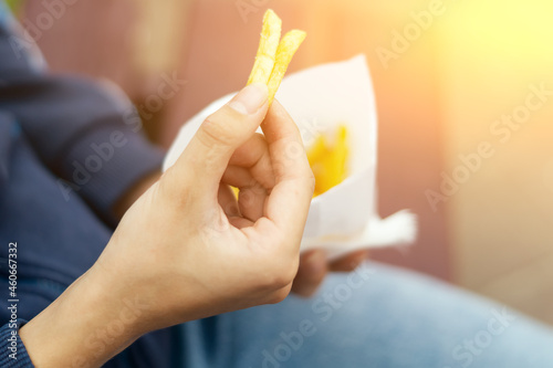 A young woman's hand takes out slices of yellow,  appetizing French fries from a white paper bag. A young girl is eating delicious French fries right on the street, sitting on a bench in a city park.