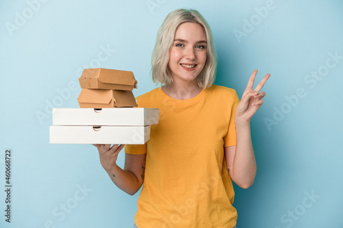 Young caucasian woman holding pizzas and burgers isolated on blue background joyful and carefree showing a peace symbol with fingers.