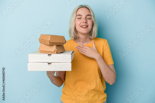 Young caucasian woman holding pizzas and burgers isolated on blue background laughs out loudly keeping hand on chest. photo