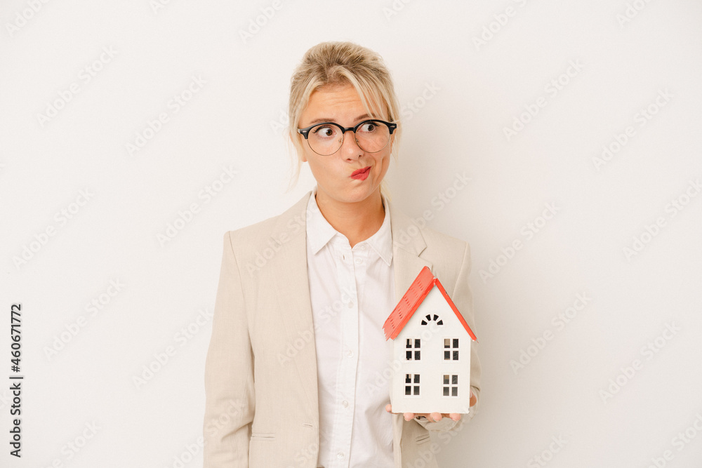 Young real estate agent woman holding a home model isolated on white background confused, feels doubtful and unsure. Young real estate agent woman holding a home model isolated on white background confused, feels doubtful and unsure.