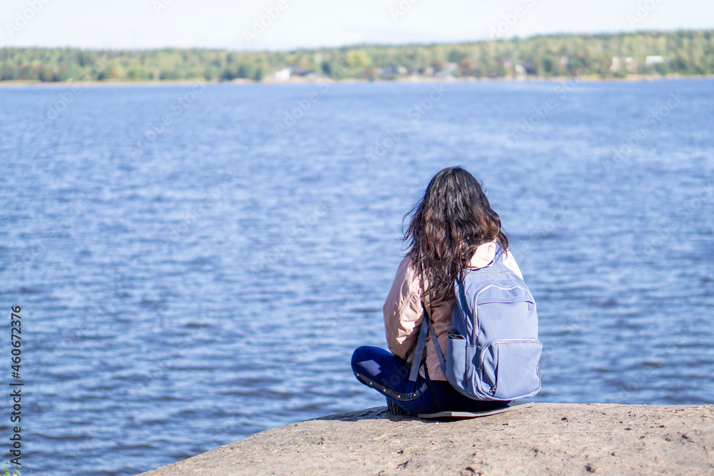 Young brunette woman sits with her back to the camera, backpack behind her shoulders and looks at the panorama of the lake. Concept of relaxation in nature.