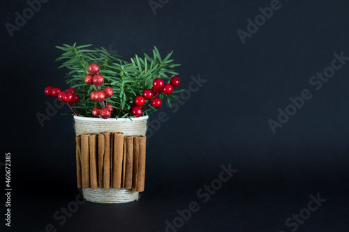 Christmas, New Year simple decor with cinnamon sticks and fir evergreen branch and red berries in a cup on black background