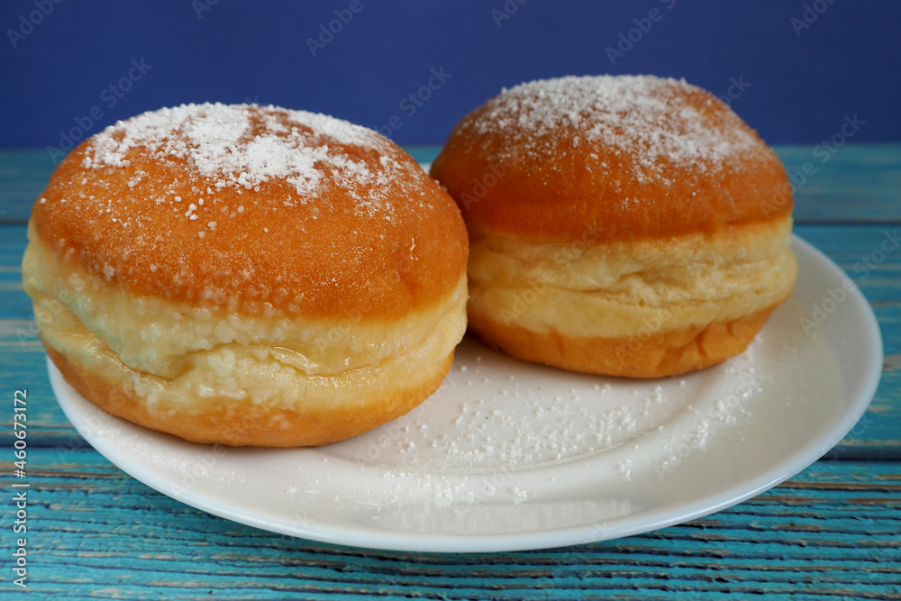 two doughnuts sprinkled with powdered sugar lie on a white plate on a blue wooden background . side view. breakfast