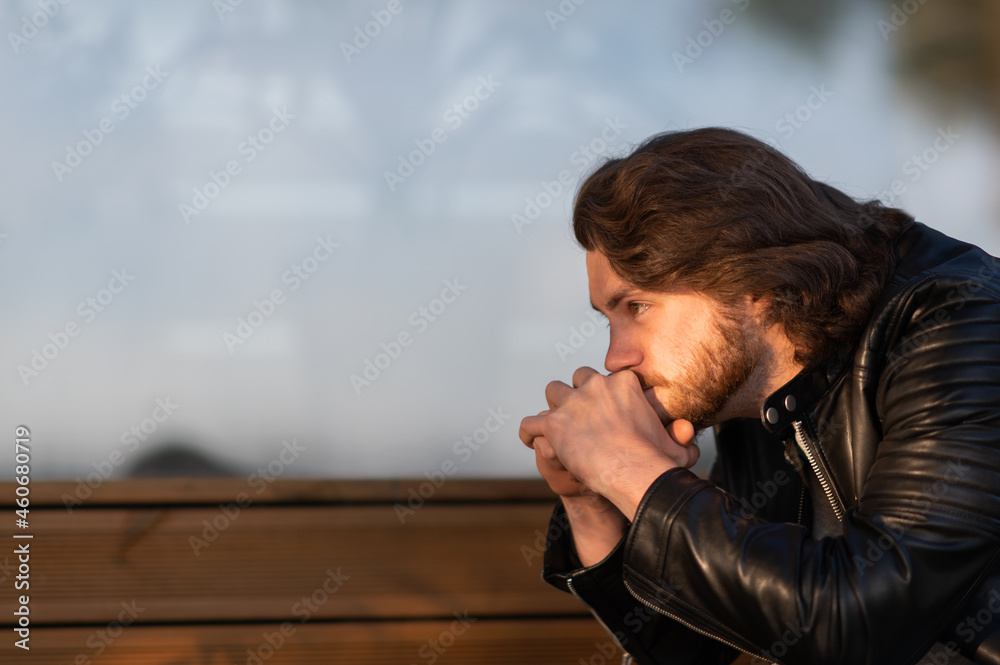 Obraz premium Pensive serious young bearded man wear black leather jacket sitting and thinking outdoors