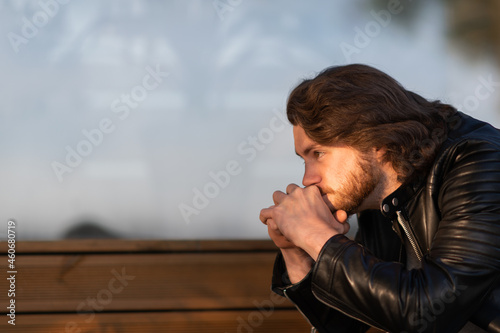 Pensive serious young bearded man wear black leather jacket sitting and thinking outdoors