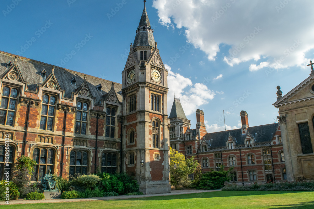View of historical bricked Christ's College building attached to a medieval style clock tower in front of lawn and benches in Cambridge England