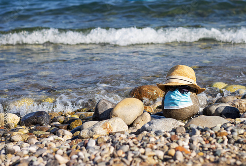 a large stone on the beach with a medical mask from coronavirus Covid-19, a hat and glasses on the background of the sea