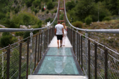 Rear view with out of focus perspective of turists walking on a suspended pedestrian bridge with steel cables in natural scenary. Italy