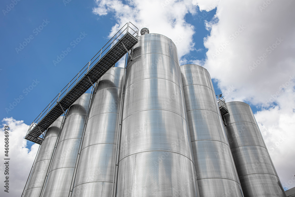 A row of stainless steel fermentation wine tanks against clouds and a blue sky. Steel wine tanks for wine fermentation at a winery. modern wine factory with large shine tanks for the fermentation. A row of stainless steel fermentation wine tanks against clouds and a blue sky. Steel wine tanks for wine fermentation at a winery. modern wine factory with large shine tanks for the fermentation.