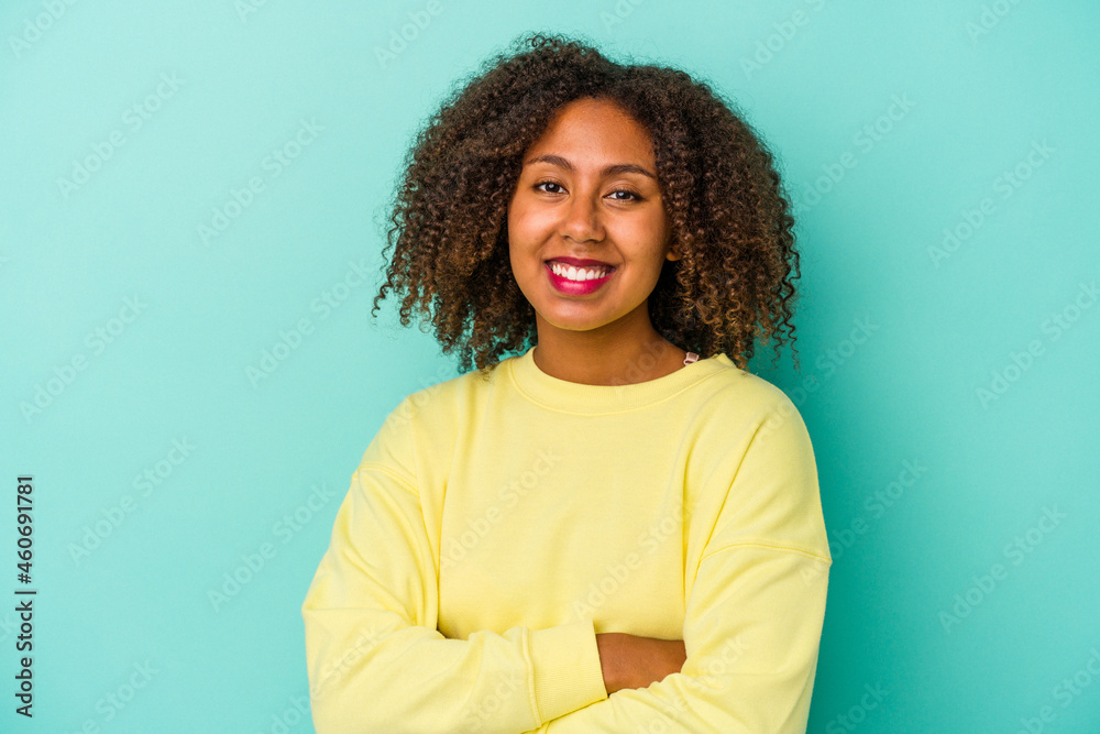 Fototapeta premium Young african american woman with curly hair isolated on blue background who feels confident, crossing arms with determination.