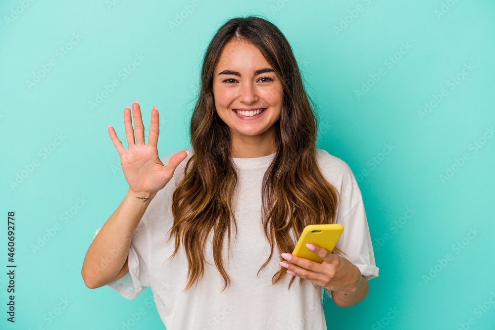 Young caucasian woman holding a mobile phone isolated on blue background smiling cheerful showing number five with fingers. Young caucasian woman holding a mobile phone isolated on blue background smiling cheerful showing number five with fingers.