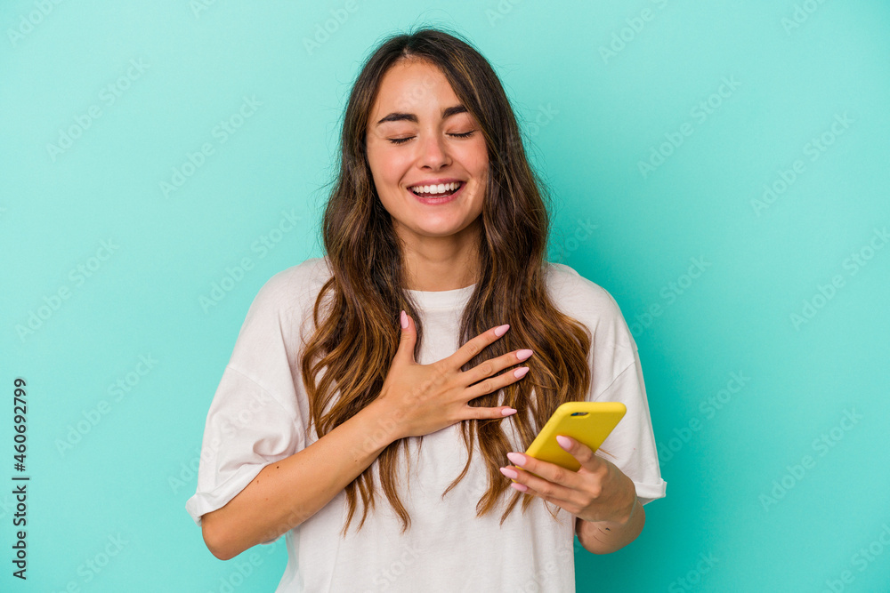Young caucasian woman holding a mobile phone isolated on blue background laughs out loudly keeping hand on chest. Young caucasian woman holding a mobile phone isolated on blue background laughs out loudly keeping hand on chest.