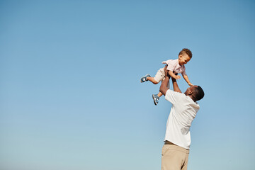 Minimal portrait of happy African-American father playing with cute baby and throwing him against blue sky, copy space