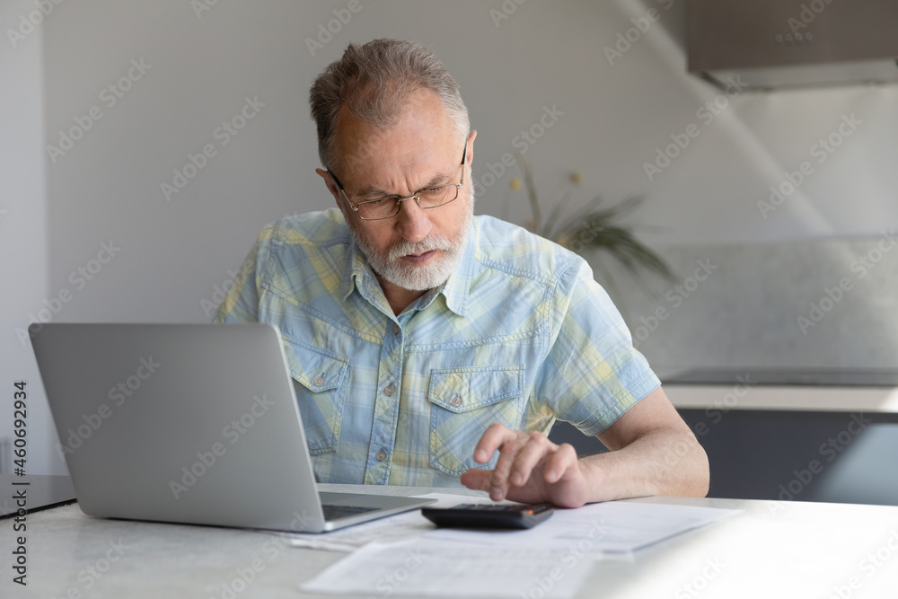 Concentrated middle aged retired man in eyeglasses using calculator, managing household budget, analyzing paper bank bills, planning investment or making online payment using computer e-banking app. Concentrated middle aged retired man in eyeglasses using calculator, managing household budget, analyzing paper bank bills, planning investment or making online payment using computer e-banking app.