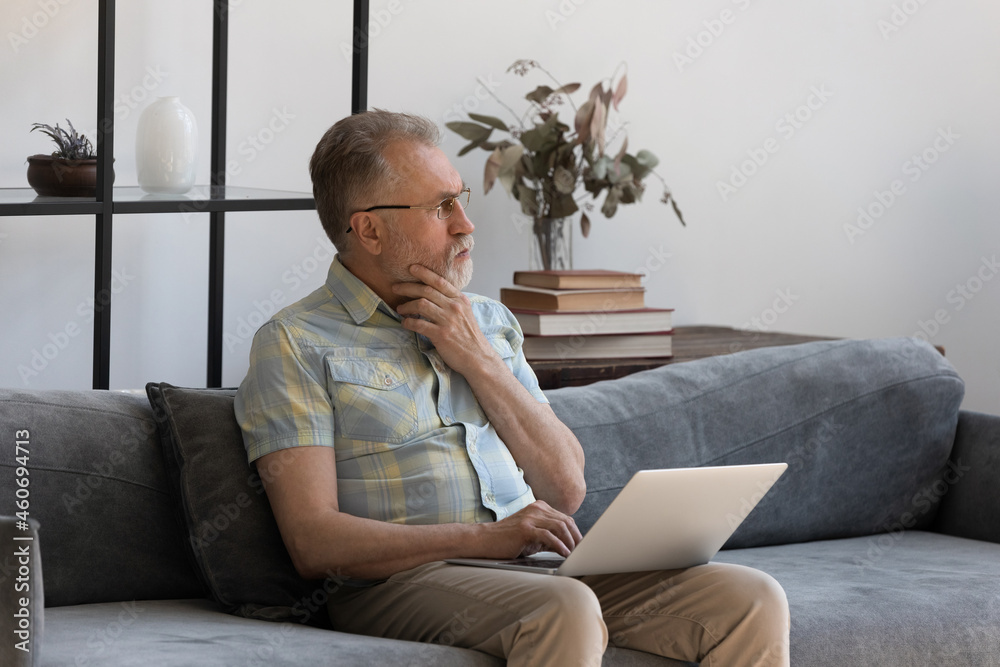 Pensive middle aged 60s man in eyewear holding computer on laps, looking in distance considering problem solution, thinking of email with bad news or having problems with modern technology gadgets.