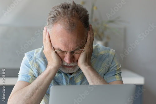 Frustrated middle aged older retied man in eyeglasses covering ears, feeling stressed using computer software applications, considering financial problem, suffering from lack of money managing budget. photo