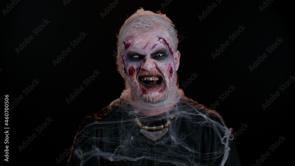 Zombie man with makeup with fake wounds scars and white contact lenses looking at camera and clicks his teeth, trying to scare in dark room. Sinister dead guy. Halloween, filming, staging concept