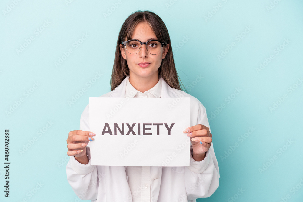 Young doctor caucasian woman holding anxiety placard isolated on blue background Young doctor caucasian woman holding anxiety placard isolated on blue background