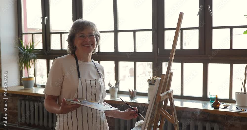 Old painter woman smiles look at camera while standing alone in modern workshop space hold palette with mixed paints and paintbrush, drawing picture on canvas on easel. Hobby, art studio class concept