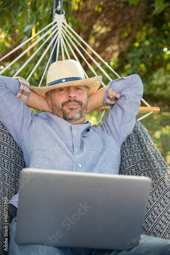 un homme d'âge mûr est allongé dans son hamac avec un chapeau de paille sur la tête, il a un ordinateur sur les genoux et il se détend, relax sous un arbre dans le jardin