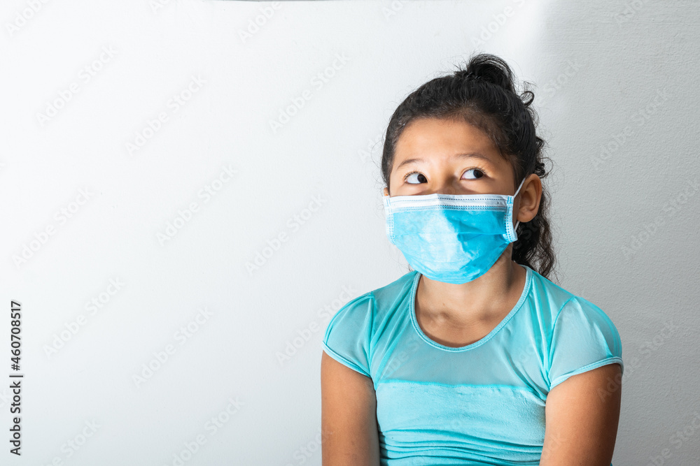 brunette girl with blue blouse and surgical mask (Latina 8 years old) pensive while looking up worried, pandemic, quarantine, covid-19. medical concept. space for advertising text