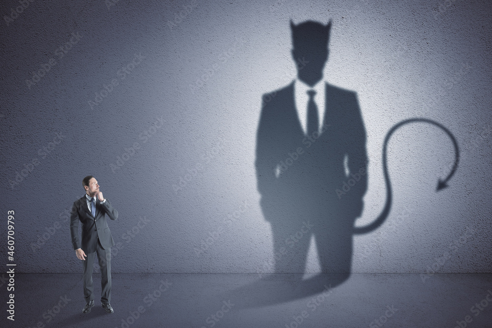 Fototapeta premium Attractive thoughtful young european male with devil shadow standing in empty concrete interior. Disguise, demon and career concept.