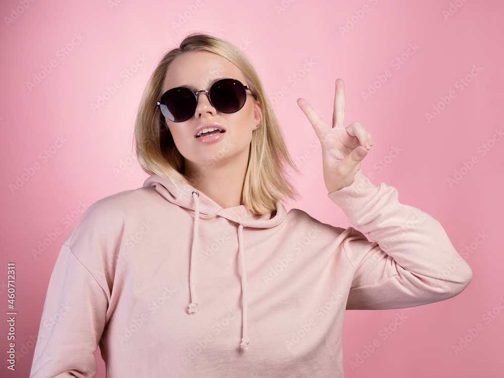 beautiful young woman with blonde hair wearing dark sunglasses makes a peace gesture, two fingers up, with one hand. In a pink hoodie on a pink background, studio portrait beautiful young woman with blonde hair wearing dark sunglasses makes a peace gesture, two fingers up, with one hand. In a pink hoodie on a pink background, studio portrait