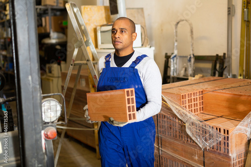 Concentrated hispanic worker stacking red bricks on pallet in warehouse of building materials ..