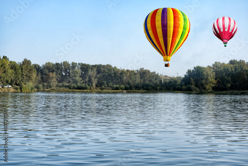 A beautiful landscape of the river surrounded by trees and hot air balloons on a sunny summer day under blue sky.