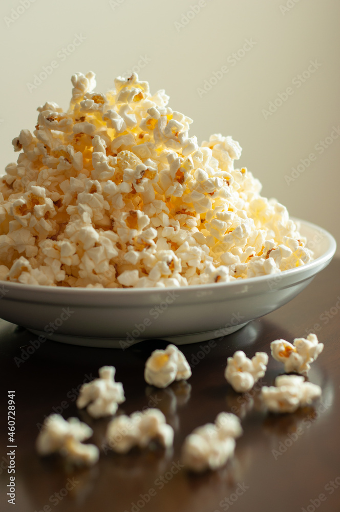 Overflowing white bowl of buttery popcorn on a dark table against a light beige wall Overflowing white bowl of buttery popcorn on a dark table against a light beige wall