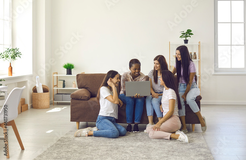 Happy friends sitting on sofa and warm heated floor in modern beautiful big apartment interior. Group of young women spending time at home, talking, using laptop computer, enjoying good movie together