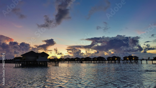 Evening twilight. There are purple and pink clouds in the blue sky. Golden glow over the horizon. Above the ocean - silhouettes of water villas. Maldives.
