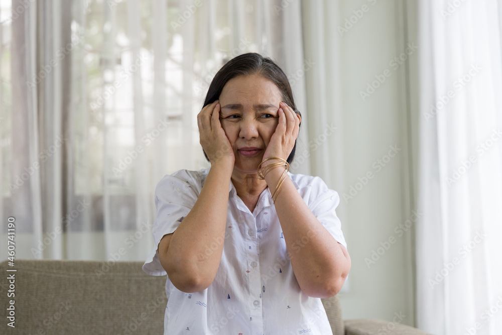 Elderly Asian woman sitting and having a headache and touching her head with her hands at home. Retirement, quarantine and health care concept Elderly Asian woman sitting and having a headache and touching her head with her hands at home. Retirement, quarantine and health care concept