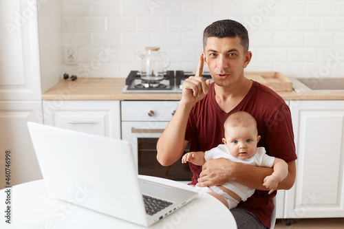 Smart brunette male wearing maroon casual style t shirt sitting at table in kitchen with his infant daughter, having clever idea, raising his finger up.