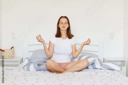 Calm relaxed female with dark hair wearing white casual style t shirt and shorts, sitting on bed in light bedroom in lotus pose, doing yoga practice and meditating.