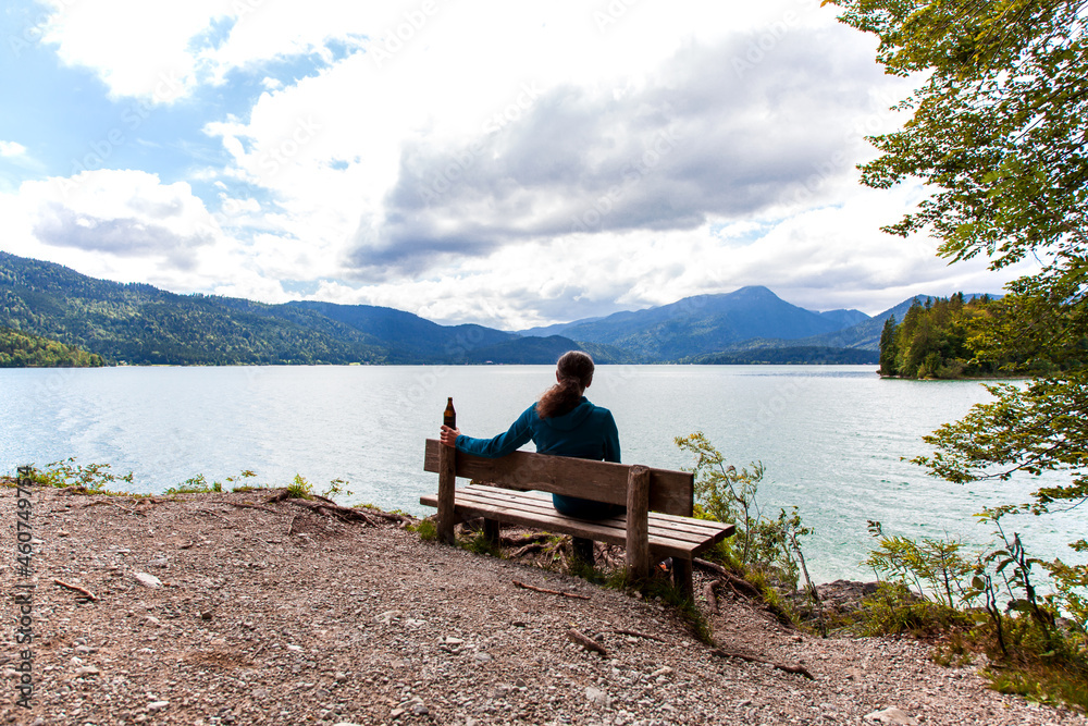 Rear view of a Man sitting on a bench for relaxation with beer bottle in green Idyllic Landscape scenery. Beautiful Bavarian panorama of lake Walchensee. Upper Bavaria Germany. Europe Bavarian Prealps