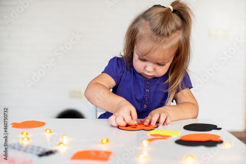 Blonde little girl created decorations for Halloween party. Cute toddler preparing for holiday Halloween. Holiday art activities, Easy felt crafts for beginners. Children's art project 