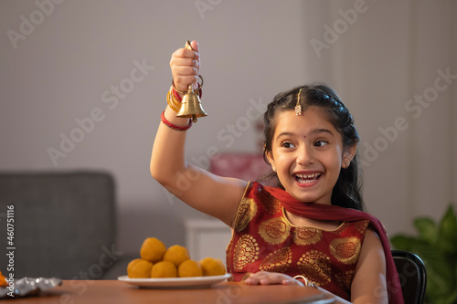 A small girl dressed In traditional cloth happily jingling a bell with a plate of ladoos kept on table. photo