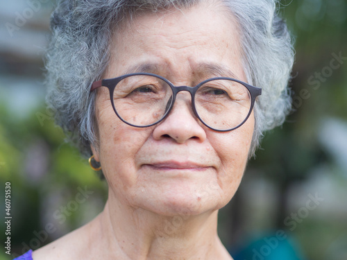 Portrait of an elderly Asian woman with short gray hair, wearing glasses, and looking away with a smile while standing in a garden. Aged people and relaxation concept