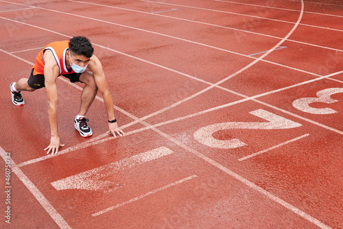 Young latin guy with protective mask, in position to run at full speed on a track, ready, at the start line, copy space