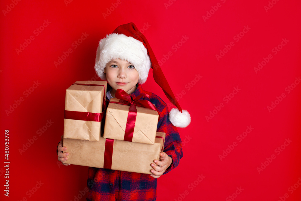 Happy holiday and Merry Christmas. Portrait of a boy in a cap with gifts in his hands on a red background. A place for text. Happy holiday and Merry Christmas. Portrait of a boy in a cap with gifts in his hands on a red background. A place for text.