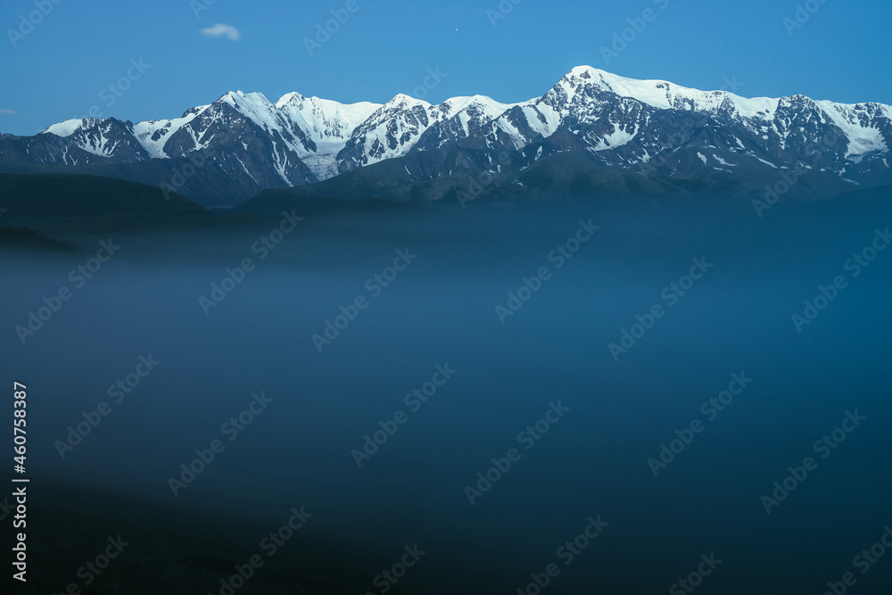 Atmospheric mountains landscape with dense fog and great snow mountain top under twilight sky. Alpine scenery with big snowy mountains over thick fog in night. High snow pinnacle above clouds in dusk.