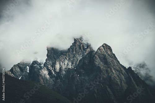 Dark atmospheric surreal landscape with dark rocky mountain top in low clouds in gray cloudy sky. Gray low cloud on high pinnacle. High black rock with snow in low clouds. Surrealist gloomy mountains. © Daniil